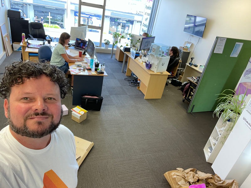 Tim in foreground of office space with Fay sitting at desk in background
